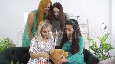 可爱的孕妇穿着蓝绿色丝绸连衣裙，向她的朋友展示泰迪熊。 四个快乐的漂亮女孩